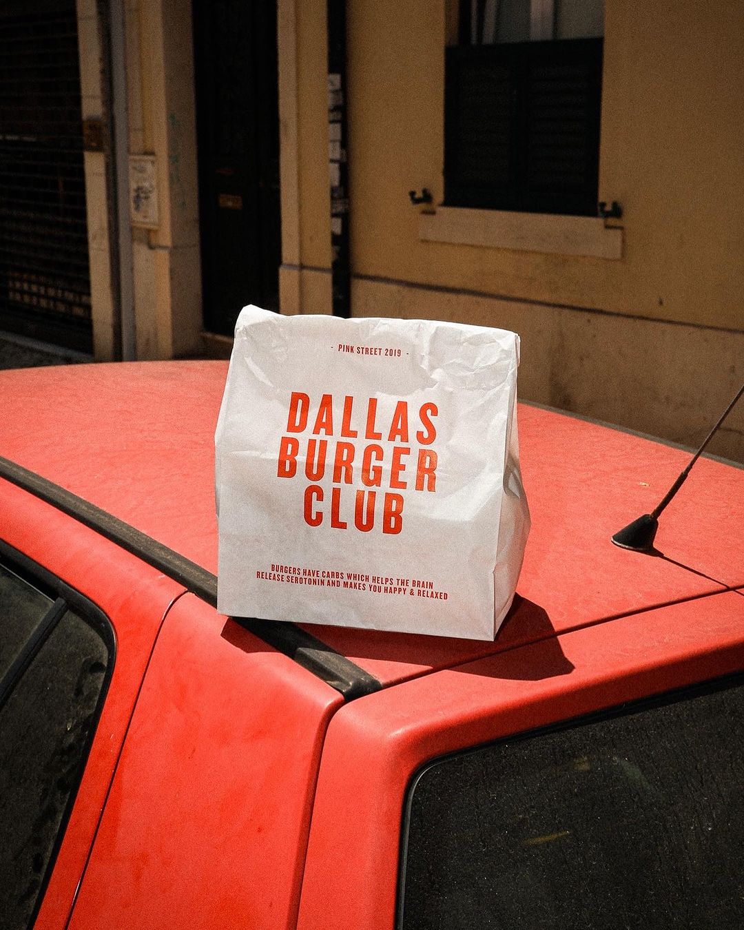 Dallas Burger Club branded paper bag without handles placed on the hood of a red car in an urban setting.
