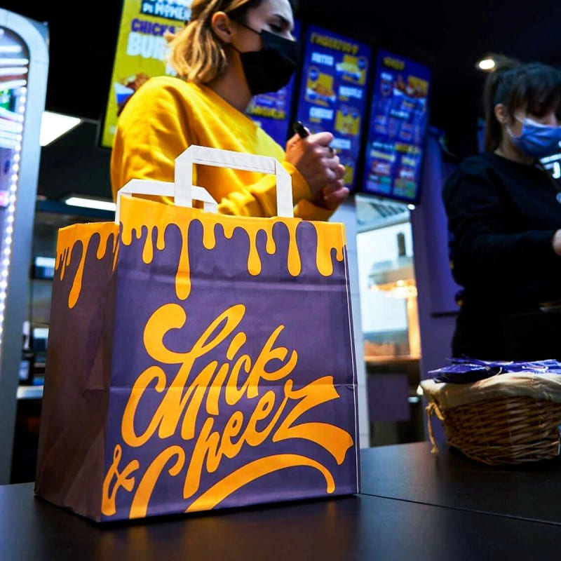 Branded takeaway bag with a vibrant design from Chick & Cheez, displayed on a counter with employees in the background.